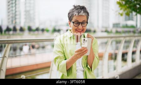 Femme mûre excitée regardant son smartphone tout en se tenant sur une passerelle moderne d'un pont urbain Banque D'Images
