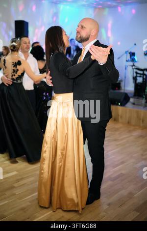 Un homme en costume sombre et cravate dorée danse avec une femme en haut noir et longue jupe dorée lors d'un bal, d'autres couples derrière eux. Banque D'Images