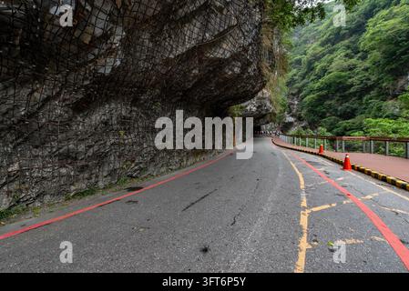 Vue du parc national de Taroko présentant une route sinueuse traversant un tunnel rocheux avec des filets de sécurité, des cônes de signalisation routière rouges et une passerelle piétonne en bois dans Banque D'Images