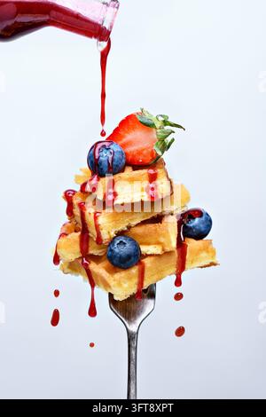 Pile de gaufres dorées garnies de fraises fraîches et de myrtilles, arrosées d'un sirop de baies rouges qui coule d'une bouteille en verre Banque D'Images