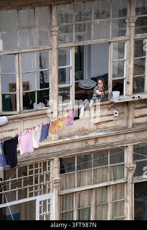Sénior accrochant du linge à la fenêtre d'une vieille maison en bois historique à Tbilissi, Géorgie. Banque D'Images