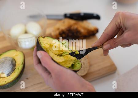 Femme adulte en manches longues claires tenant une moitié d'avocat avec une cuillère noire sur une planche à découper en bois Banque D'Images