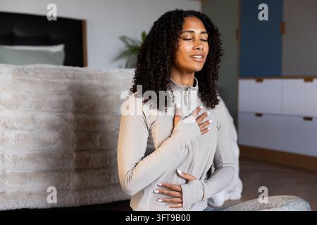 Femme afro-américaine assise sur le sol de la chambre en train de méditer dans un haut gris à fermeture éclair près du lit Banque D'Images