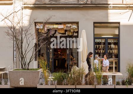 Une entrée de café de rue charmante avec une terrasse extérieure dotée de plantes en pot, un homme et une femme en conversation, et une exposition décorative fantaisiste de linge Banque D'Images