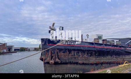 Wolfsburg, Basse-Saxe, Allemagne, Europe : L'étrave sombre du navire Nabonid est amarré au rivage. L'ancre, la corde d'amarrage, la bitture et les marques d'usure évidentes sur la coque sont visibles. Allemagne, Europe : Le bateau fluvial Nabonid est amarré avec des câbles d'amarrage à une digue dans un bassin portuaire. Le gros plan latéral montre la coque noire partiellement rouillée avec l'ancre, le nom du navire et les installations techniques. À l'arrière-plan, on distingue des surfaces d'eau et des bâtiments industriels sous un ciel densément nuageux. L'image documente la navigation fluviale, l'infrastructure portuaire et d Banque D'Images