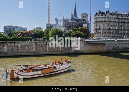 Une péniche néerlandaise traditionnelle arborant le drapeau des Pays-Bas transporte des fans en orange le long de la Seine, avec la flèche reconstruite de la cathédrale Notre-Dame et ses grues se dressant en arrière-plan à Paris, France, le 5 août 2024, pendant les Jeux Olympiques de Paris 2024. Banque D'Images