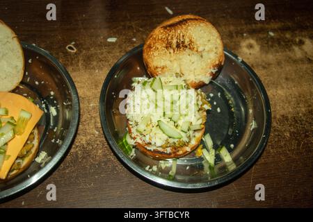 Deux sandwichs ouverts sur des assiettes en métal garnis de fromage râpé blanc et de salade de concombre en dés. Assemblage de plats de street food sur une table sombre. Banque D'Images