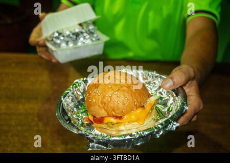 Close up d'une main tenant un délicieux cheeseburger végétarien enveloppé dans du papier aluminium brillant. Parfait pour la livraison de street food, menu d'entreprise de restauration rapide Banque D'Images