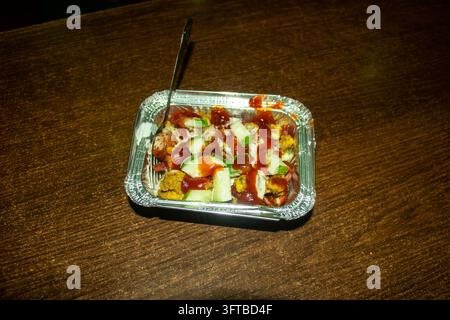 Petit plateau rectangulaire en aluminium unique contenant des morceaux de nourriture savoureuse hachés garnis de cubes de concombre frais et de ketchup tomate rouge sur une table en bois. Banque D'Images
