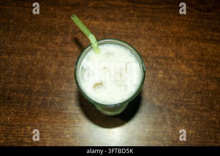 Prise en hauteur d'un milkshake blanc rafraîchissant servi dans un verre sur une table en bois brun, mettant en valeur sa texture crémeuse avec des glaçons visibles par-dessus. Banque D'Images