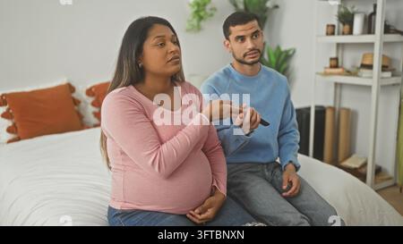 Femme enceinte tenant une télécommande sur le lit, main sur le ventre, partenaire gesticulant avec la main ouverte à côté d'elle ; tension relationnelle. Banque D'Images
