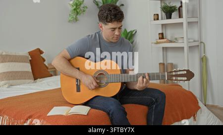 Un homme est assis sur un lit tenant une guitare acoustique, il joue et écrit dans un carnet tout en regardant vers le bas dans une chambre ; concentration créative. Banque D'Images