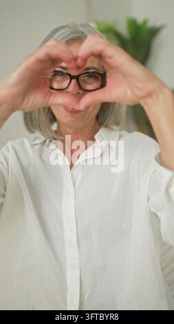 Sénior âgée avec lunettes sourit et forme un cœur avec ses mains dans un studio, assise près d'une plante ; affection douce. Banque D'Images
