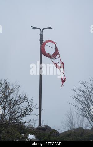 MATEUR DE VENT ROUGE DÉCHIRÉ DANS LE BRUME, ÎLE FINLANDAISE ÉLOIGNÉE EN HIVER PROFOND, UTÖ, FINLANDE : Un mateur de vent rouge déchiré et en lambeaux pend mollement à un poteau métallique, son tissu déchiqueté et détruit, sous un ciel morne et brumeux. INFO : Utö est une île finlandaise éloignée dans la mer des Archipels, au sud de Turku. Connue pour son phare historique, ses paysages côtiers accidentés et son héritage maritime, c’est l’île habitée la plus méridionale de Finlande, attirant les visiteurs en quête de tranquillité, d’observation des oiseaux et de paysages spectaculaires de la mer Baltique. Banque D'Images