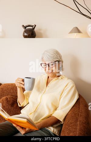 Sénior femme profitant de son temps libre sur un canapé, lisant un livre et tenant une tasse de café Banque D'Images