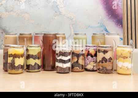 Bocaux de dessert superposés variés avec brownie, crème anglaise, crème à la pistache et aux fruits rouges ainsi que café glacé, thé et boissons au lait dans des boîtes transparentes avec des cuillères sur un lig Banque D'Images