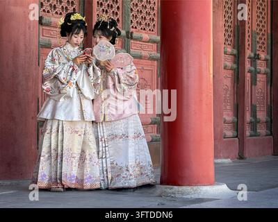 Deux femmes en robe traditionnelle chinoise regardant un smartphone à l'intérieur de la Cité interdite à Pékin, en Chine, à côté de murs et de colonnes rouges ornés. Banque D'Images