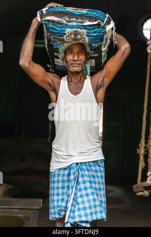 Un porteur vêtu d'un lungi bleu à carreaux soulève un sac lourd au-dessus de sa tête à deux mains dans une salle de stockage sombre, Bengale occidentale, Howrah, Inde Banque D'Images