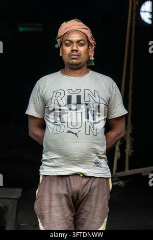 Portrait frontal d'un ouvrier en short marron et t-shirt imprimé dans un entrepôt sombre avec des cordes derrière lui, Bengale occidentale, Howrah, Inde Banque D'Images