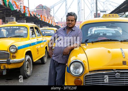 Un homme en chemise bleue s'appuie nonchalamment sur le capot d'un taxi jaune classique tandis que d'autres taxis s'alignent sous un pont, Bengale occidentale, Kolkata, Inde Banque D'Images
