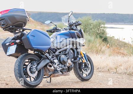 Moto de tourisme bleue avec des sacoches et une valise sur le toit, garée sur un chemin de terre à côté d’un réservoir en Estrémadure, en Espagne, avec des collines et de la végétation en arrière-plan Banque D'Images