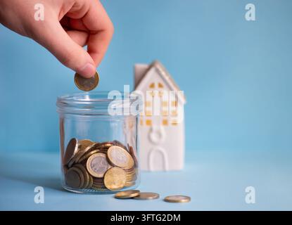 Main mettant une pièce en euro dans un pot d'épargne en verre avec une maison miniature sur fond bleu. Concept d'épargne domestique et de budget ménager. Banque D'Images