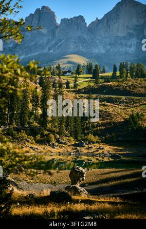 Prairie alpine baignée de soleil avec un lac de montagne réfléchissant, une ferme sur le milieu de la colline et des pics imposants des Dolomites en arrière-plan, une lumière dorée du soir illuminant Banque D'Images