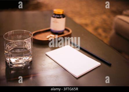Un verre d'eau clair repose à côté d'un bloc-notes vierge, d'un stylo noir, d'une bouteille de médicaments et de comprimés sur une table sombre à l'intérieur Banque D'Images