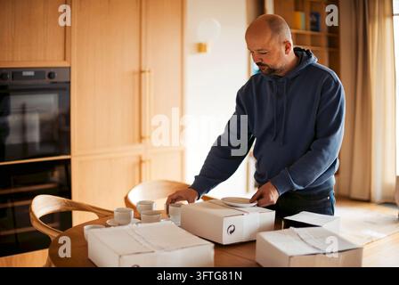Homme chauve portant un sweat à capuche bleu marine range des emballages en carton blanc à côté de gobelets sur une table en bois, avec des placards de cuisine, des appareils électroménagers et des fenêtres derrière lui Banque D'Images