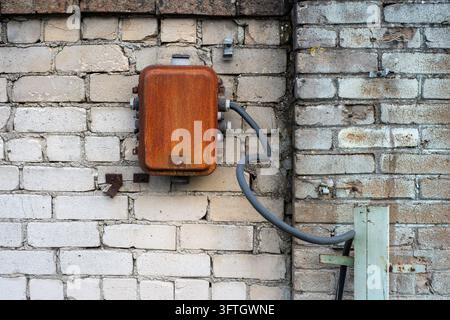 Ancienne boîte de jonction électrique rouillée avec câble exposé monté sur un mur de briques délavé dans un cadre industriel. Banque D'Images