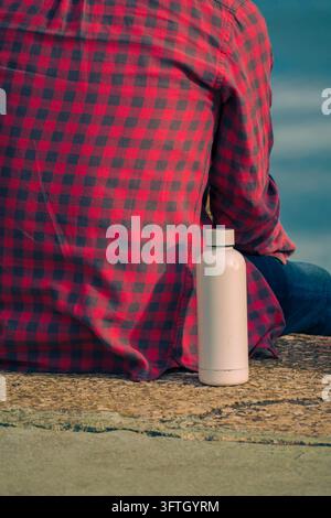 Homme en chemise à carreaux rouge assis en contemplation à côté d'une bouteille d'eau près de l'eau Banque D'Images