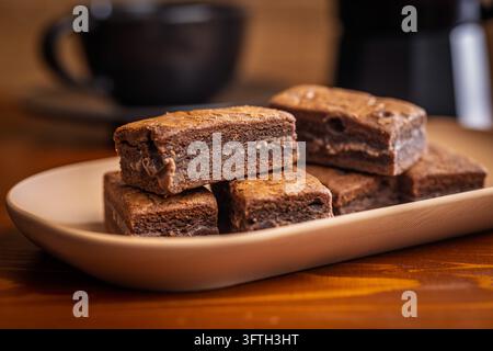 Brownies au chocolat fondants et empilés servis sur une assiette beige avec une tasse de café floue en arrière-plan. Photographie culinaire chaleureuse et confortable mettant en valeur la texture Banque D'Images