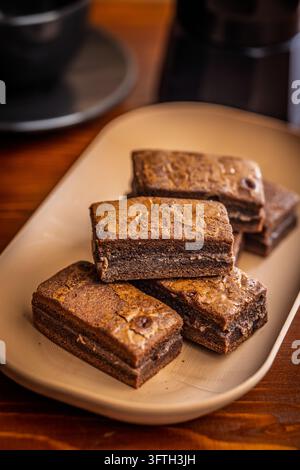 Brownies au chocolat fondants et empilés servis sur une assiette beige avec une tasse de café floue en arrière-plan. Photographie culinaire chaleureuse et confortable mettant en valeur la texture Banque D'Images