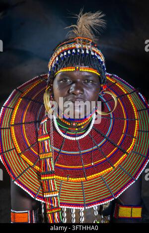 Femme du tribe Pokot dans une hutte sombre avec un couvre-chef en plumes, des boucles d’oreilles en laiton et un collier de perles décoré de coquilles, Comté de Baringo, Churo, Kenya Banque D'Images