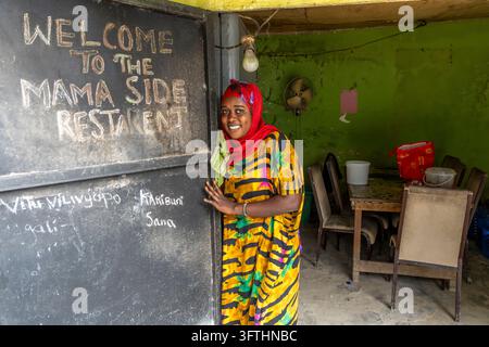 Femme souriante portant un hijab rouge et une robe à motifs jaunes accueillant à l'entrée du restaurant Mama Side, Stone Town, Tanzanie Banque D'Images