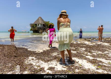 Touristes en robes colorées posant et prenant des photos sur une plage couverte d'algues devant le restaurant The Rock à Zanzibar, Jambiani, Tanzanie Banque D'Images