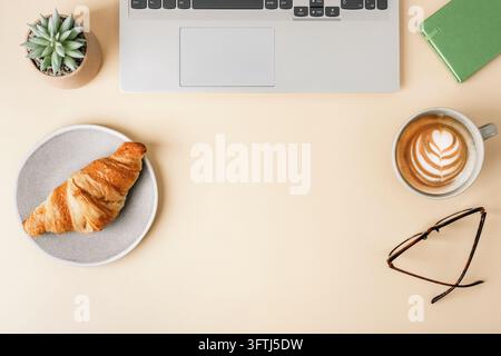 Ordinateur portable, croissant, cappuccino, succulent et lunettes sur un bureau beige avec espace de copie. Vue de dessus, flat lay. Banque D'Images