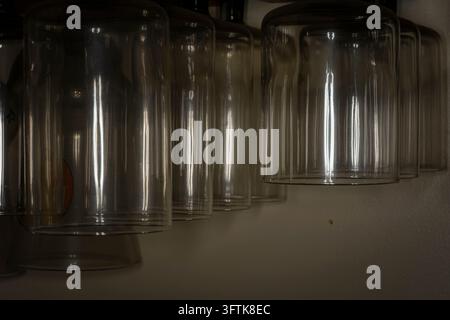 Range de verres à boire clairs suspendus à l'envers sous une lumière faible et atmosphérique dans un cadre de cuisine ou de bar Banque D'Images
