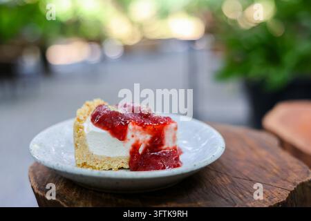 Une part de cheesecake avec garniture de fraises sur une assiette en céramique, photographiée en plein air avec un feuillage vert flou et une table en bois rustique créant une ambiance détendue Banque D'Images