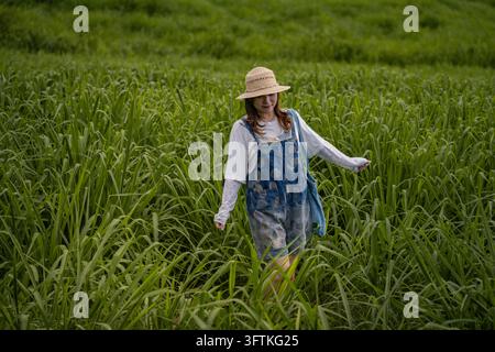 Jeune femme souriante en salopette en jean marchant dans une haute herbe verte en touchant les brins, Nghia Dan, Nghe An, Vietnam Banque D'Images