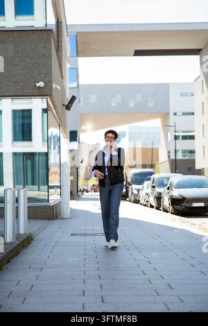 Une femme d'affaires mûre souriante parle au téléphone tout en tenant un ordinateur portable dans une rue de Helsinki devant un bureau moderne Banque D'Images