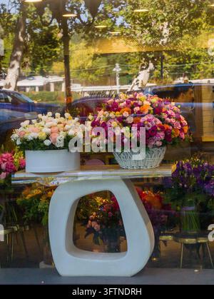 Arrangements floraux vibrants exposés dans une vitrine de magasin. Des fleurs blanches et colorées sont posées sur une table blanche moderne. Les réflexions de voitures et d'arbres apparaissent sur Banque D'Images