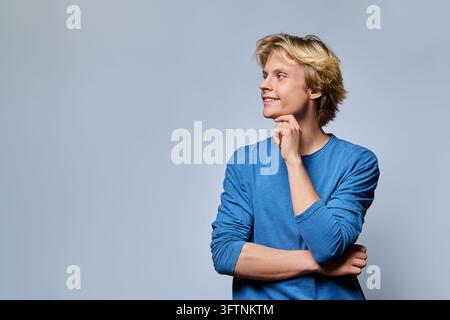Vue de côté d'un jeune homme blond souriant regardant l'espace de copie dans le studio. Concept d'idées créatives, pensées d'étudiant, vision future, curiosité, inspiration Banque D'Images