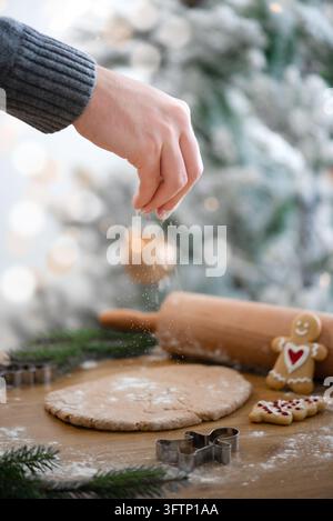 Main saupoudrant de la farine sur de la pâte à pain d'épices roulée sur une table en bois avec un rouleau à pâtisserie, des emporte-pièces et des biscuits de Noël décorés. Banque D'Images