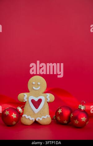 Homme en pain d'épice souriant avec un cœur rouge et un glaçage blanc, entouré de trois boules de Noël rouges avec des étoiles dorées sur un tissu rouge avec espace libre. Banque D'Images