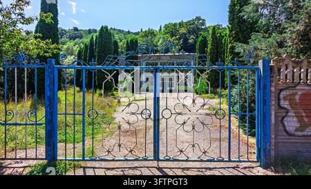 Une porte métallique bleue est ouverte devant une maison rustique. De grands cyprès et des arbustes verts entourent la propriété. La lumière du soleil projette des ombres sur le vieux Banque D'Images