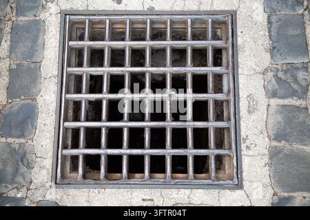 Grille d'évacuation métallique carrée encastrée dans un pavé fissuré avec une surface érodée, une ouverture sombre en dessous et des textures de maçonnerie rugueuses, vue de dessus en milieu urbain Banque D'Images