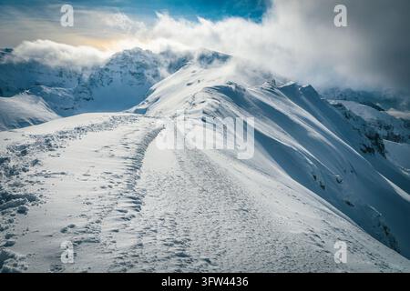 Une vue panoramique spectaculaire d'une crête montagneuse enneigée avec des nuages bas dérivant au-dessus des sommets accidentés et des empreintes de pas creusées dans la poudreuse profonde et gelée Banque D'Images