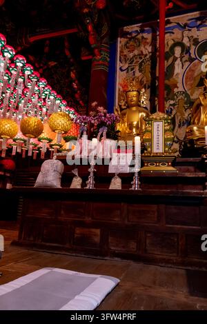 Une vue latérale à l'intérieur d'un temple montre une statue dorée, des autels en bois avec des bougies blanches, des sacs d'offrandes et des rangées de lanternes en papier colorées lumineuses suspendues Banque D'Images