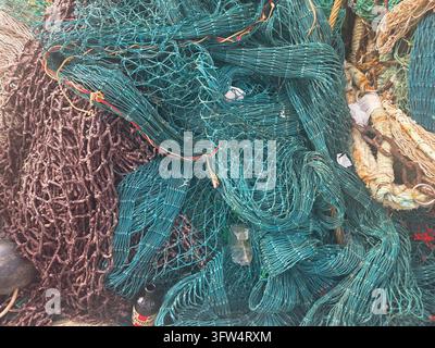 Grand tas enchevêtré de filets de pêche commerciaux bleu-vert et marron avec des cordes et des chaînes empilés sur un quai de pêche. Banque D'Images